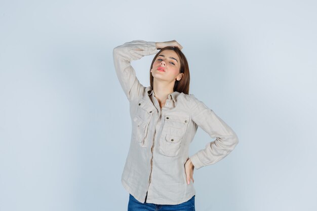 Young girl holding one hand on head, another hand on hip in beige shirt, jeans and looking pretty. front view.