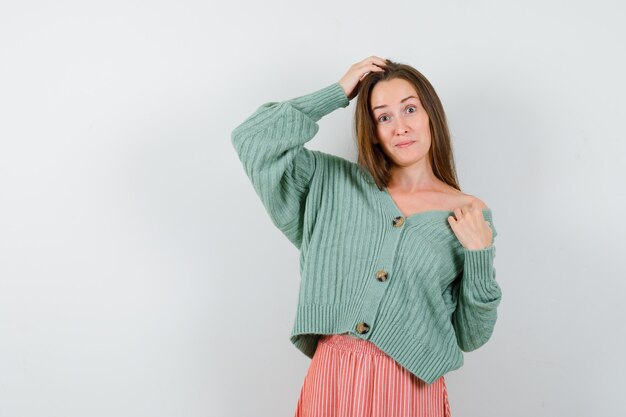 Young girl holding one hand on head, another hand on shoulder in knitwear, skirt and looking serious. front view.