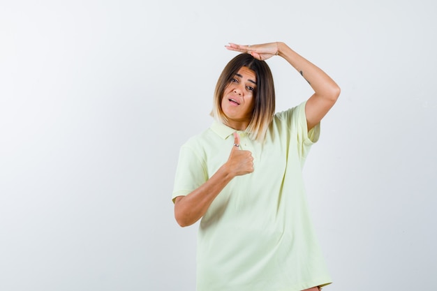 Young girl holding one hand on head, showing thumb up in t-shirt and looking happy , front view.