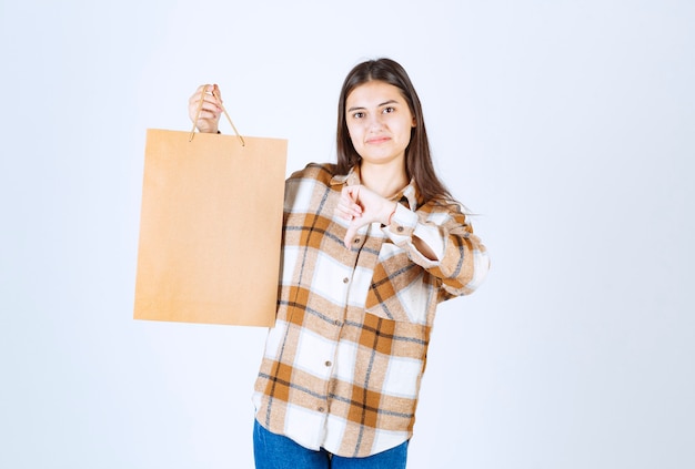 Young girl holding paper craft package and giving thumbs down over white wall. 