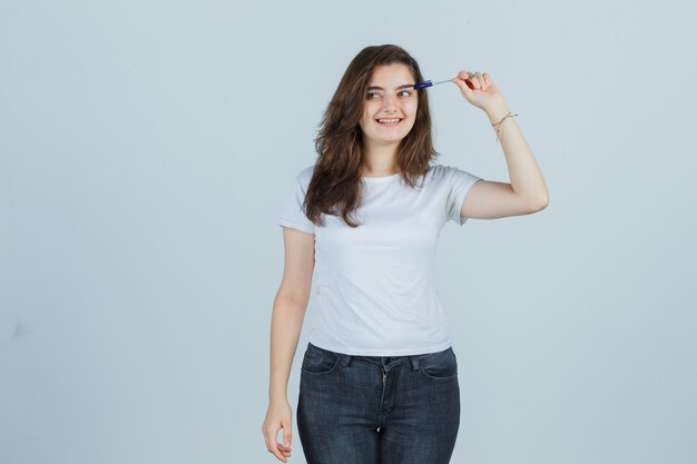 Young girl holding pen on head in t-shirt, jeans and looking joyful. front view.