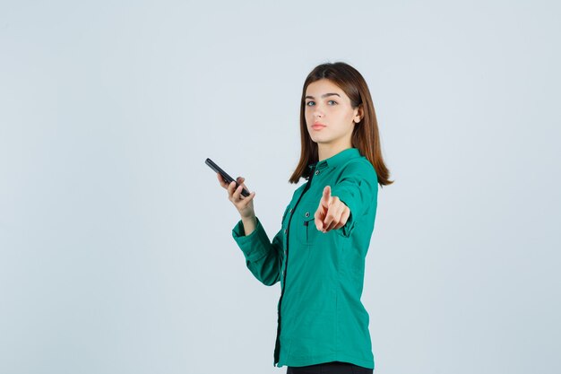 Young girl holding phone in one hand, pointing at camera in green blouse, black pants and looking serious. front view.