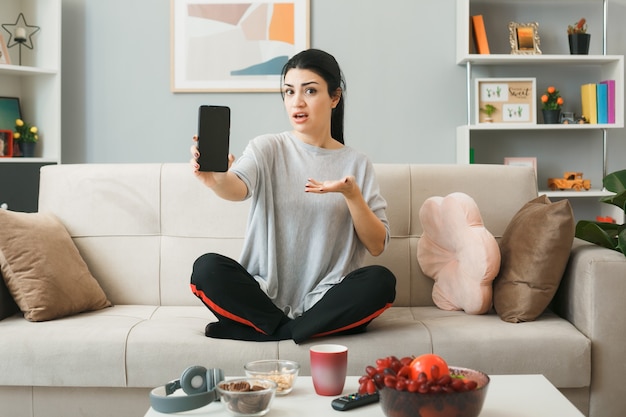 Young girl holding and points with hand at phone sitting on sofa behind coffee table in living room