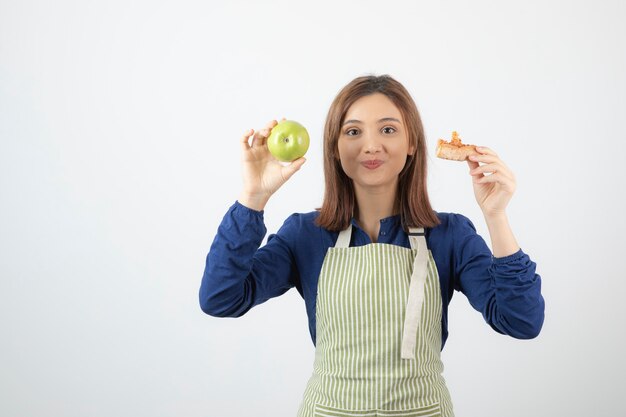 young girl holding slice of pizza and apple on white wall.