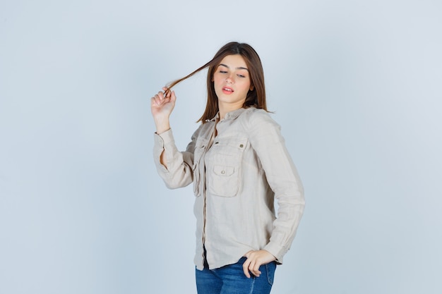 young girl holding strand of hair, with hand on hip while posing in beige shirt, jeans and looking attractive. front view.