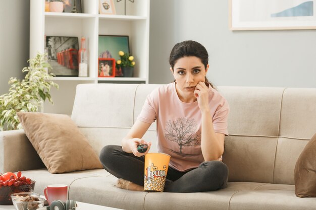Молодая девушка держит пульт от телевизора, сидя на диване за журнальным столиком в гостиной