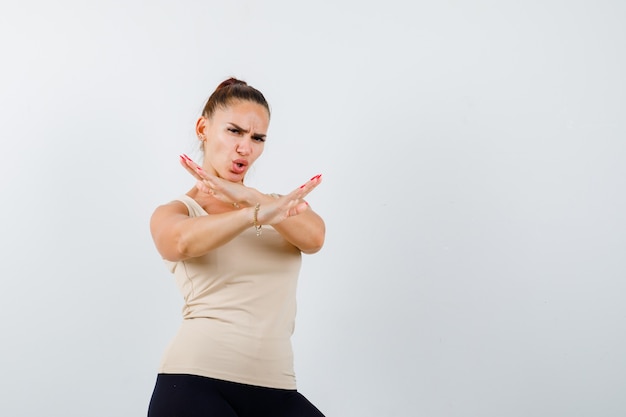 Free photo young girl holding two arms crossed, gesturing no sign in beige top, black pants and looking angry , front view.