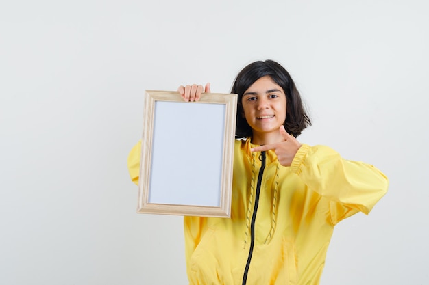 Young girl holding whiteboard and pointing to it with index finger in yellow bomber jacket and looking happy