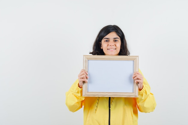 Free photo young girl holding whiteboard with both hands in yellow bomber jacket and looking happy.