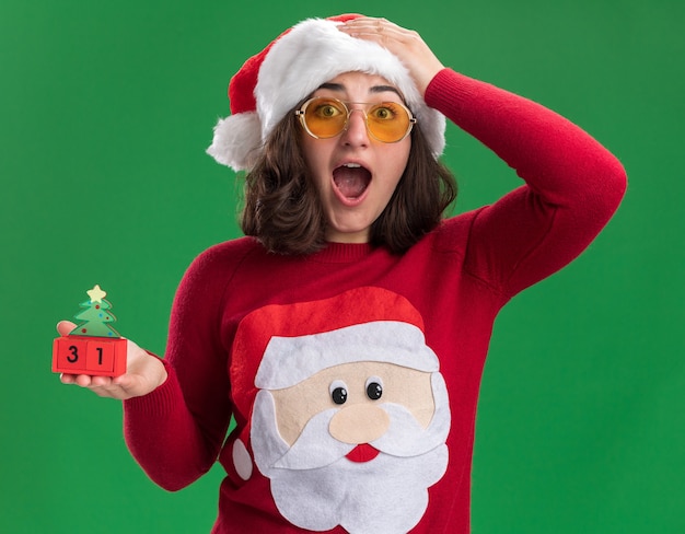 無料写真 サンタの帽子と新年の日付でおもちゃの立方体を保持しているメガネを身に着けているクリスマスセーターの少女は、緑の壁の上に立って驚いて驚いた