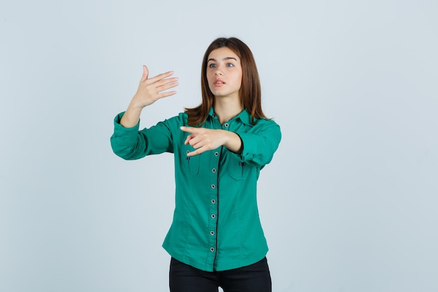 Бесплатное фото Молодая девушка в зеленой блузке, черных штанах, протягивающая руку, держащая что-то воображаемое, демонстрирующая рок-н-ролльный жест и сосредоточенная, вид спереди.