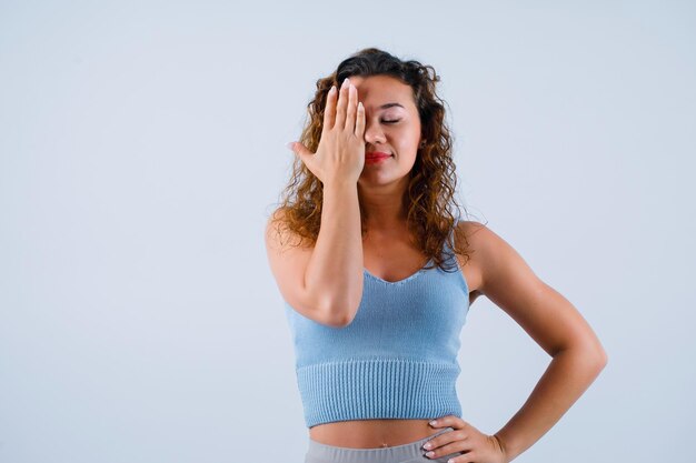 Young girl is covered left side of face with hand and putting other hand on waist on white background