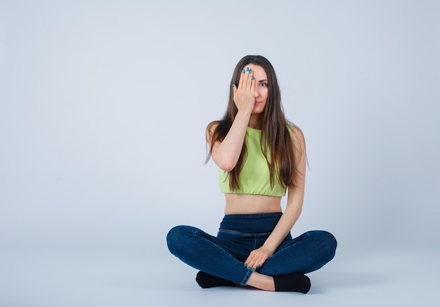 Free photo young girl is covering her half face with hand by sitting on floor on white background