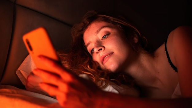 若い女の子はベッドの中で彼女のスマートフォンにいます。部屋の赤い照明