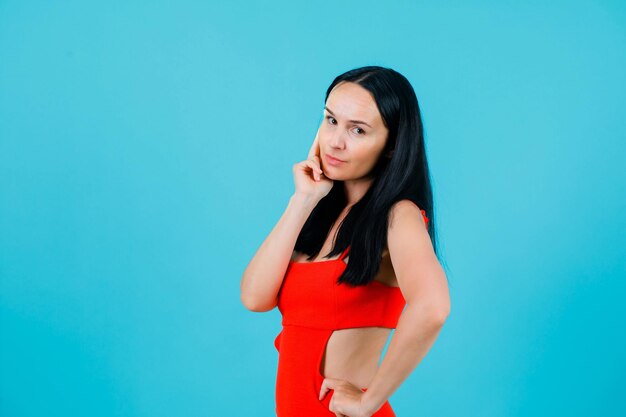 Young girl is looking at camera by holding hand on cheek and putting other hand on waist on blue background