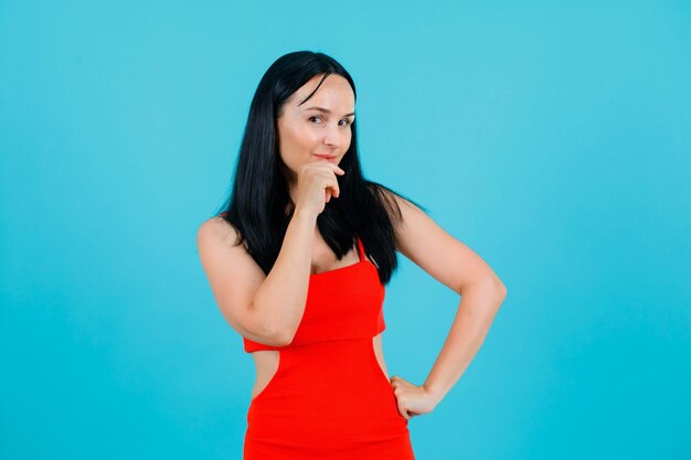 Young girl is looking at camera by holding hand on chin an dputting other hand on waist on blue background