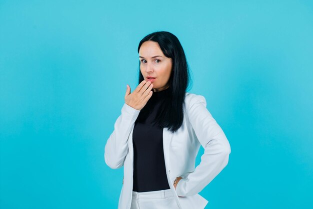 Young girl is looking at camrea by holding hand on mouth and putting other on waist on blue background