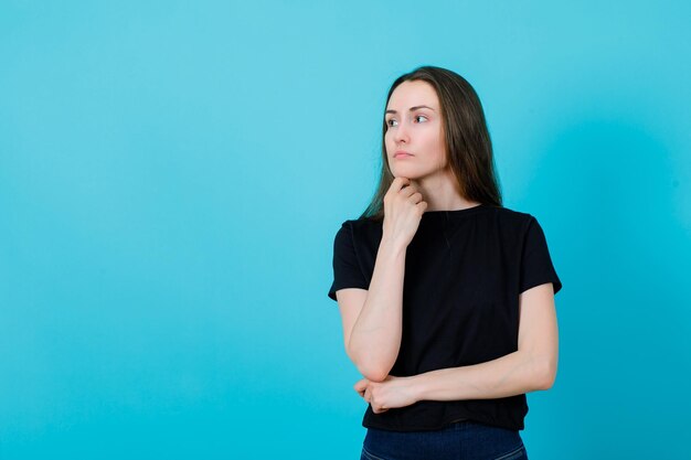 Young girl is looking at left by holding hand under chin on blue background