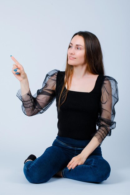 Young girl is looking at left a nd pointing left with forefinger by sitting on floor on white background