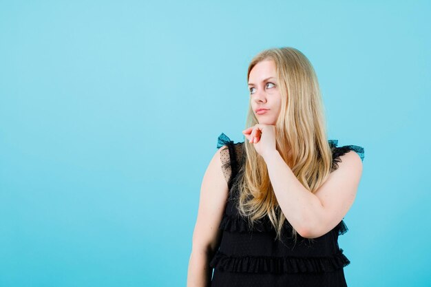 Young girl is looking left up by holding hand under chin on blue background