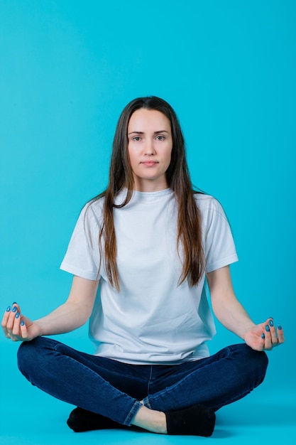 Young girl is meditating by sitting on floor on blue background