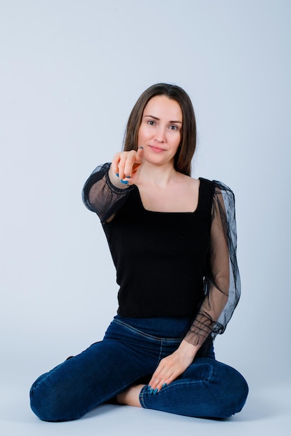 Young girl is pointing to camera with forefinger by sitting on floor on white background