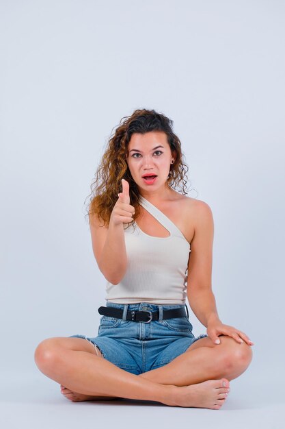Young girl is pointing to camera with forefinger by sitting on floor on white background