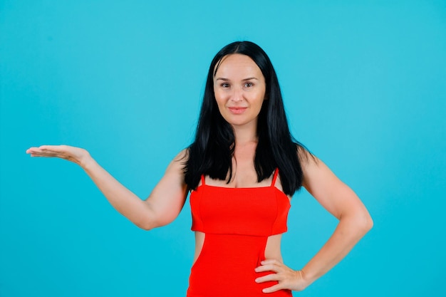 Young girl is pointing left with hand and putting other hand on waist on blue background