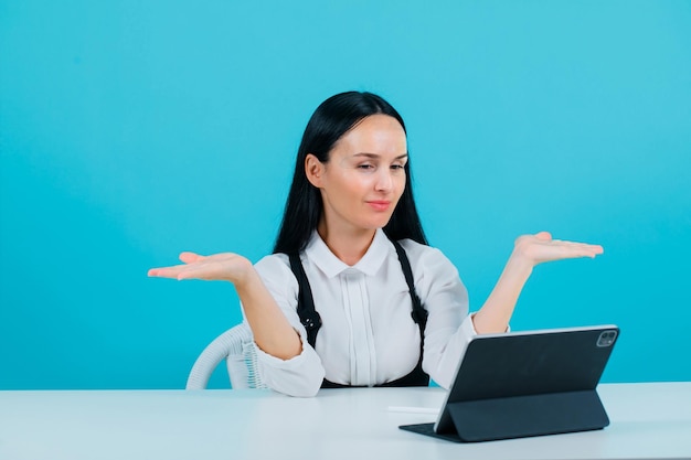 Young girl is posing to tablet camera by opening wide her hands on blue background