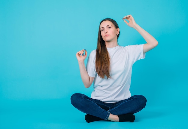 Young girl is raising up he rhands by sitting on floor on blue background