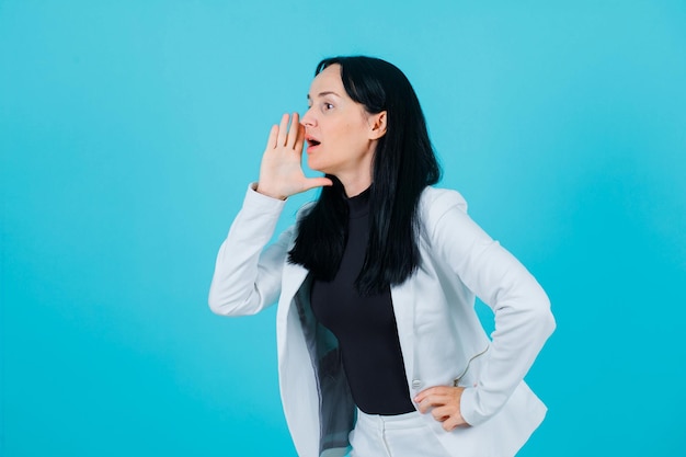 Young girl is screaming by holding ahnd near mouth and putting other on waist on blue background