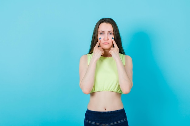 Young girl is showing crying gesture by putting forefingers on cheeks on blue background