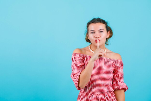 Young girl is showing silence gesture by holding forefinger on lips on blue background