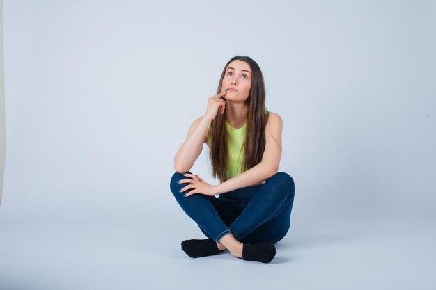 Young girl is sitting on floor and thinking by holding forefinger on chin on white background