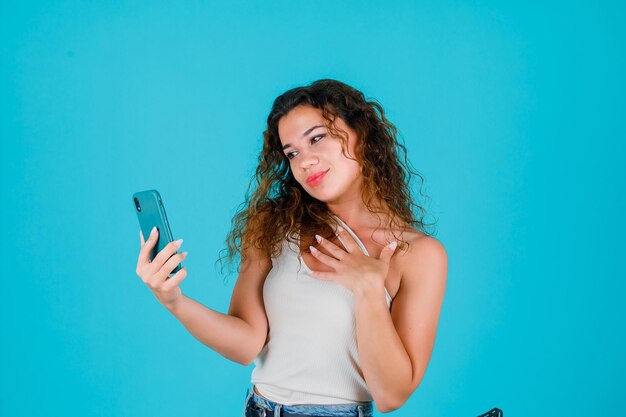 Young girl is taking selfie by holding hand on chest on blue background