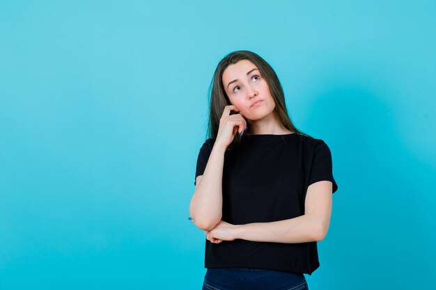 Young girl is thinking by holding forefinger on cheek on blue background