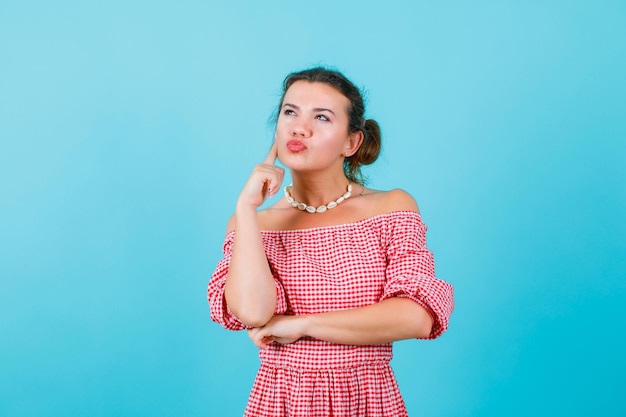 Young girl is thinking by holding forefinger on cheek on blue background