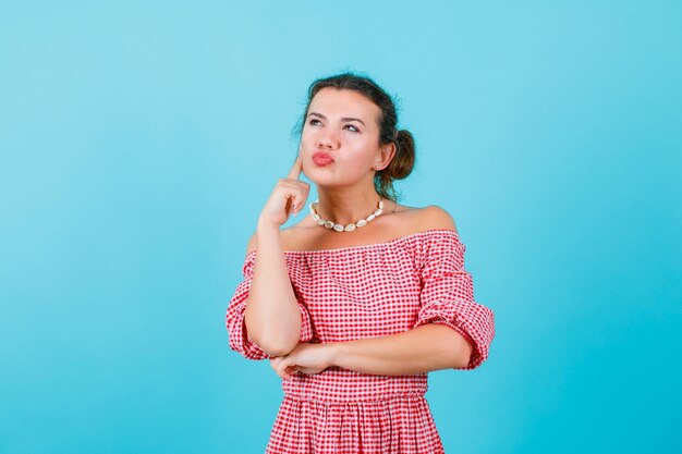 Young girl is thinking by holding forefinger on cheek on blue background