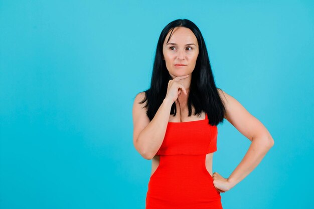 Young girl is thinking by holding hand under chin and putting other hand on waist on blue background