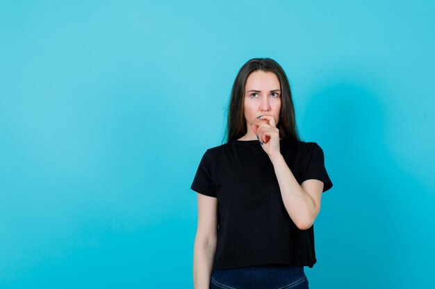 Young girl is thinking by putting hand on chin on blue background