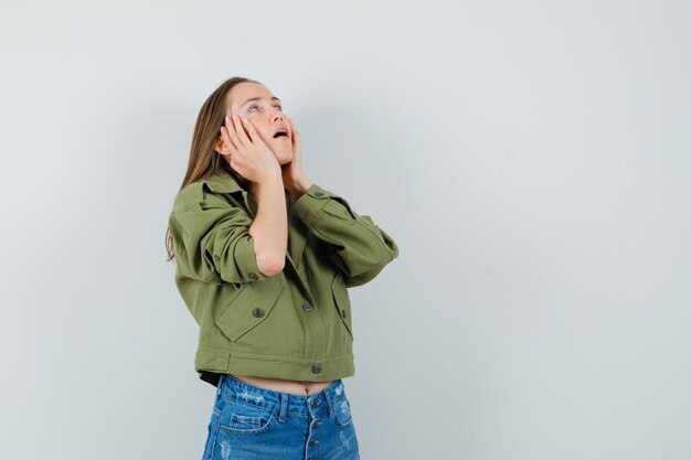 Young girl in jacket, shorts holding hands on cheeks and looking tired , front view.