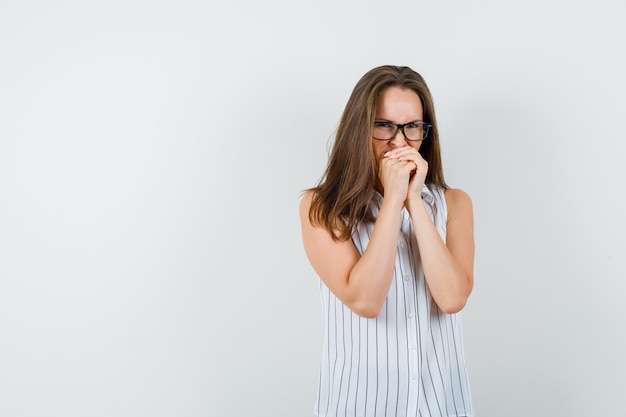 Young girl keeping clasped hands on mouth in t-shirt and looking scared , front view.