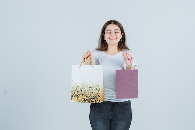 Young girl keeping gift bags in t-shirt, jeans and looking pleased , front view.