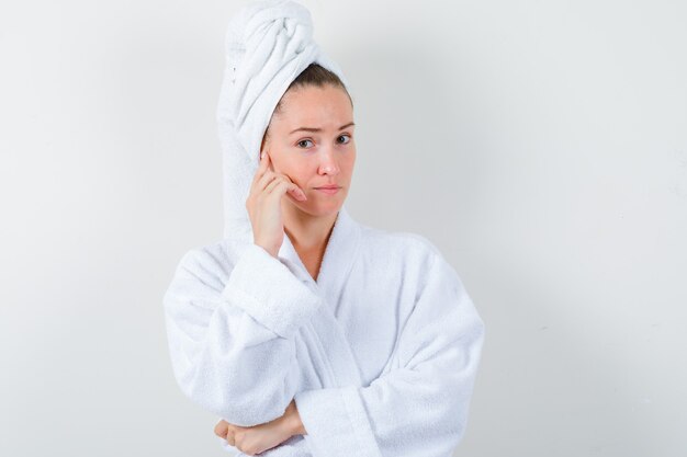 Young girl keeping hand on cheek in white bathrobe, towel and looking pensive , front view.