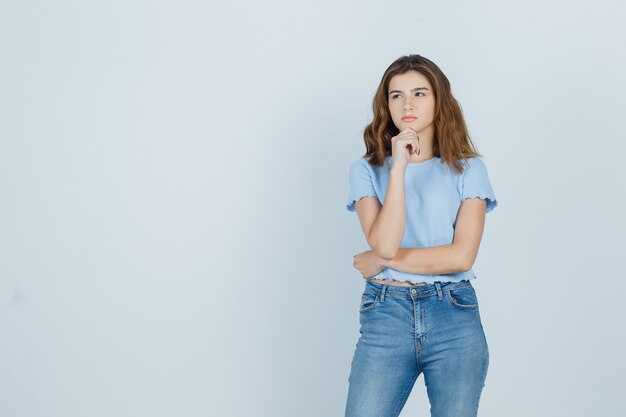 Молодая девушка держит руку на подбородке в футболке, джинсах и смотрит вдумчиво, вид спереди.