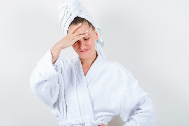 Young girl keeping hand over head in white bathrobe, towel and looking sorrowful. front view.