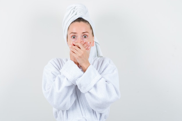 Young girl keeping hands on mouth in white bathrobe, towel and looking scared , front view.