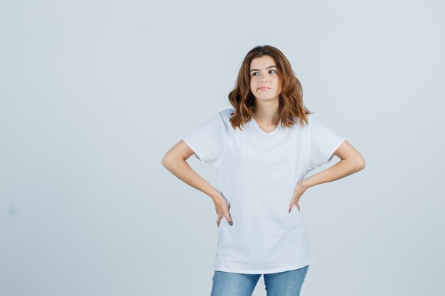 Young girl keeping hands on waist in white t-shirt and looking disappointed. front view.