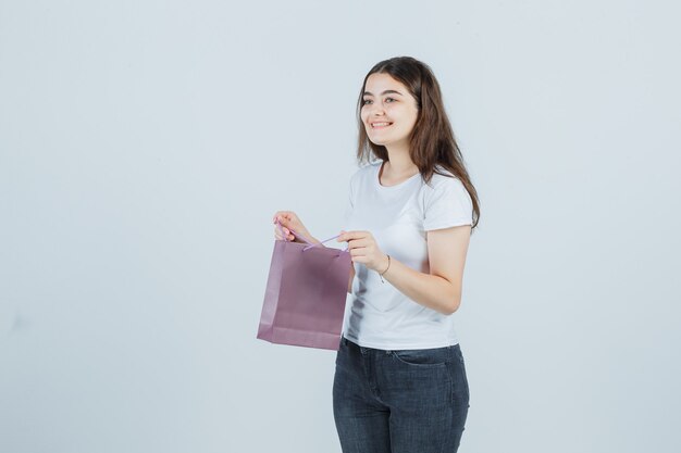Молодая девушка держит бумажный пакет в футболке, джинсах и выглядит веселой, вид спереди.