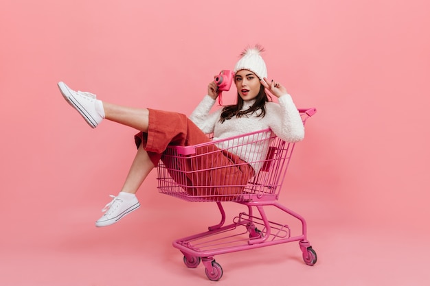 Young girl in knitted clothes poses with pink camera while sitting in supermarket trolley on isolated wall.
